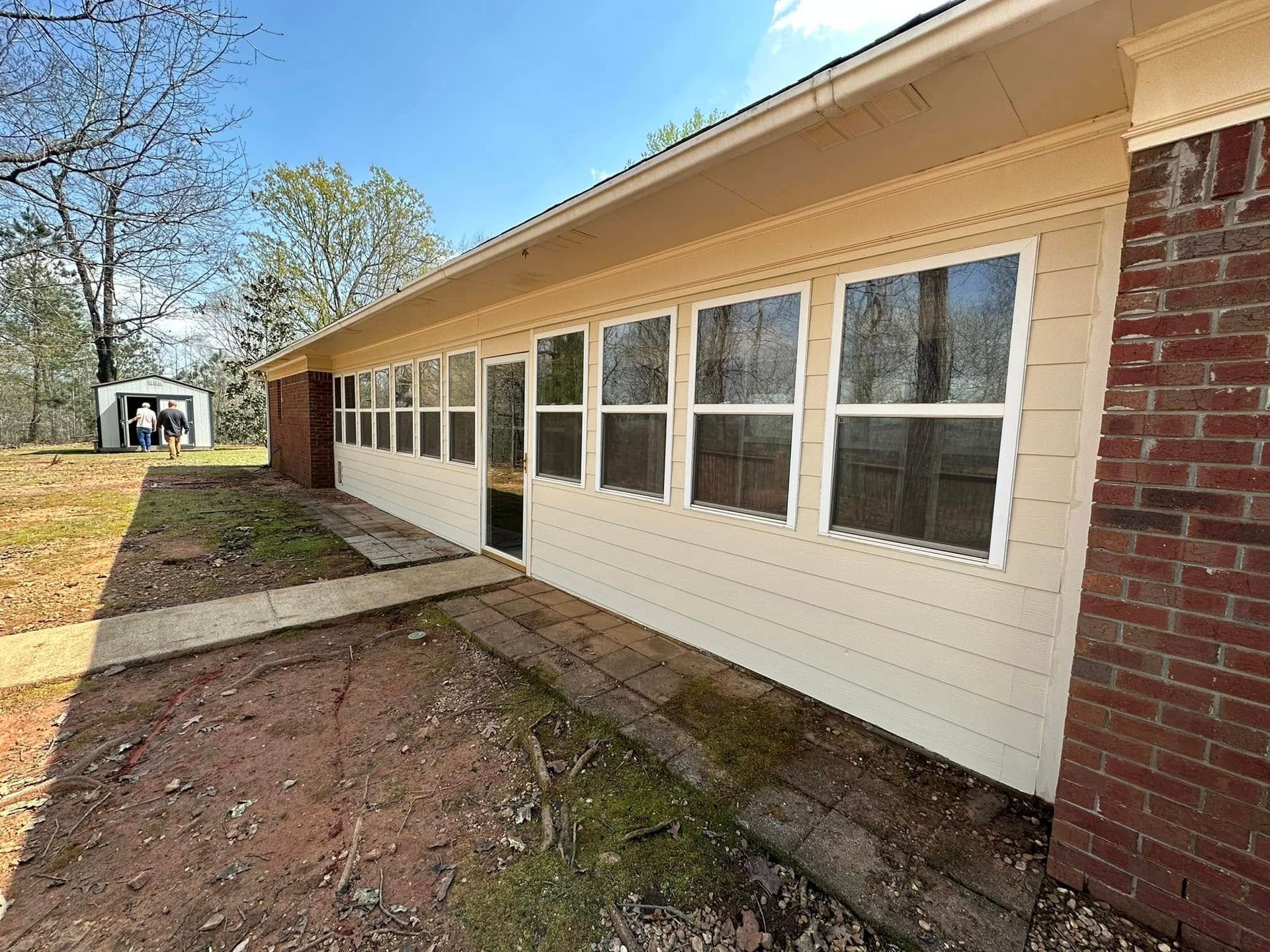 Single-story beige building with large windows, brick corner, and dirt path alongside it under a blue sky