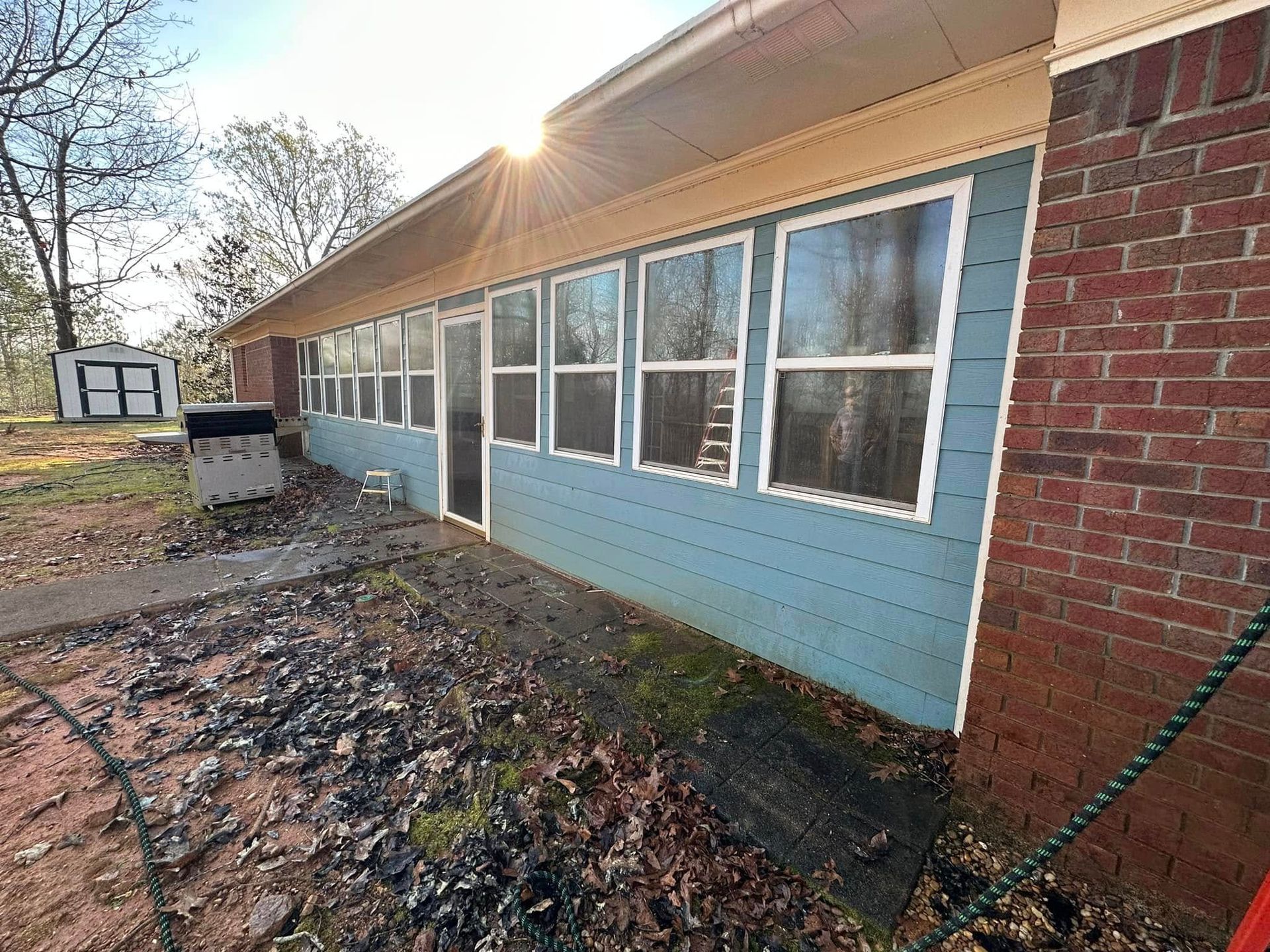Blue house exterior with large windows and red brick corner, viewed from a leaf-covered yard