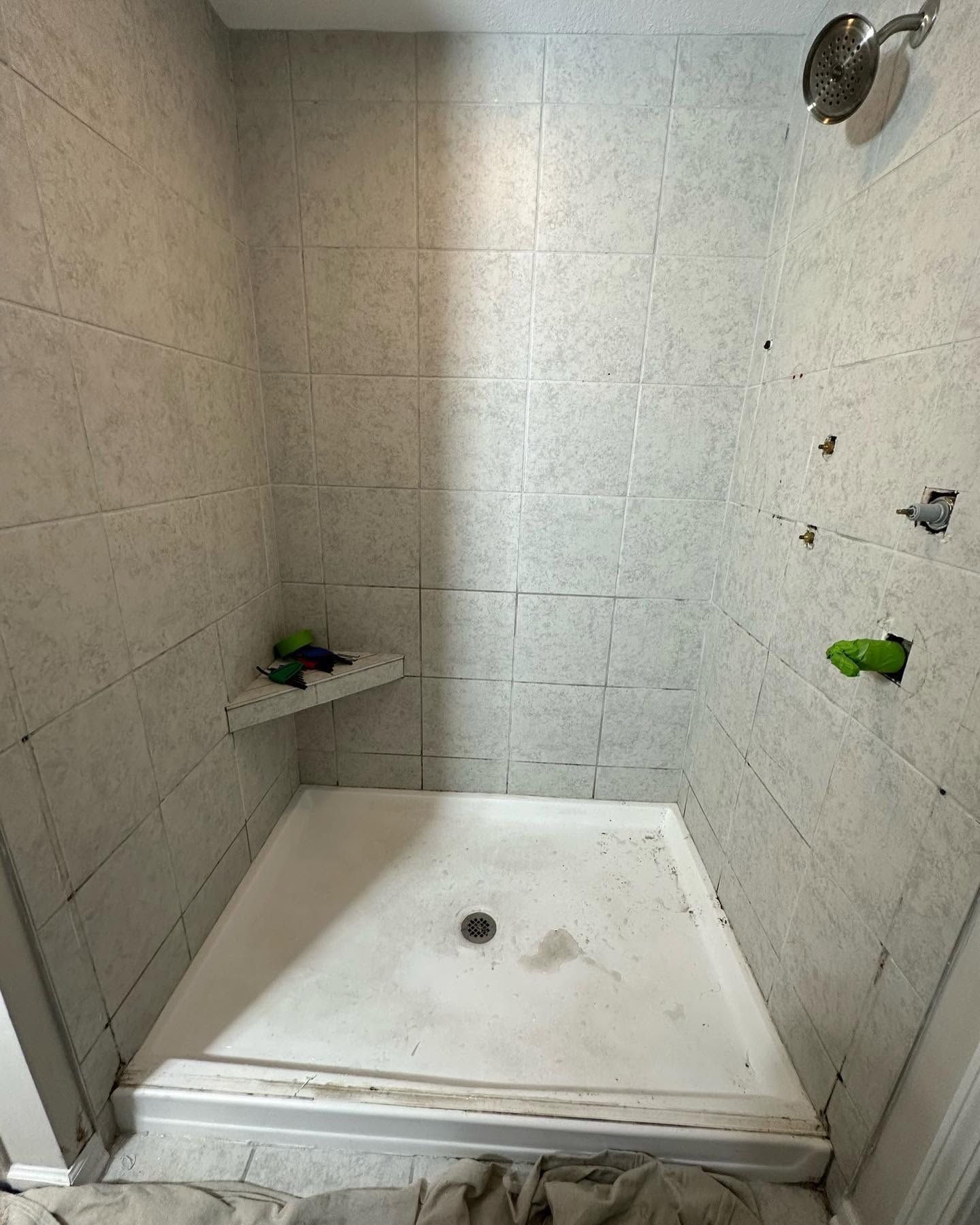 Small tiled shower stall with a white base, wall shelf, and showerhead.