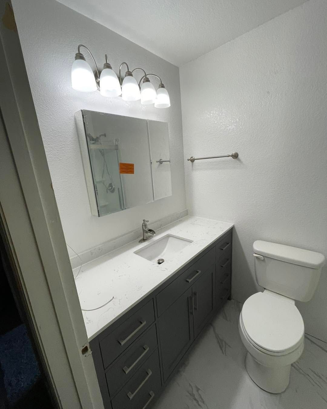Small white bathroom with vanity, mirror, lights, towel bar, and toilet.