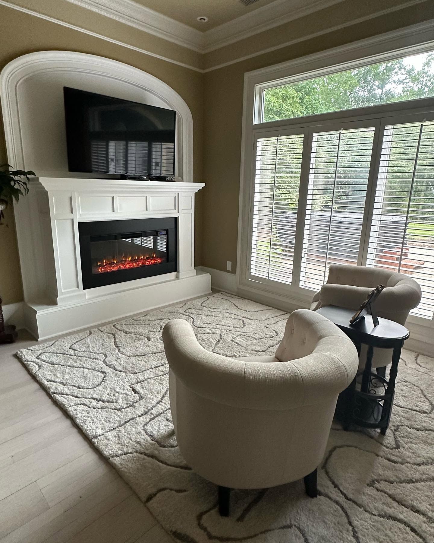 Cozy living room with white fireplace, beige chairs, patterned rug, and large window with blinds.