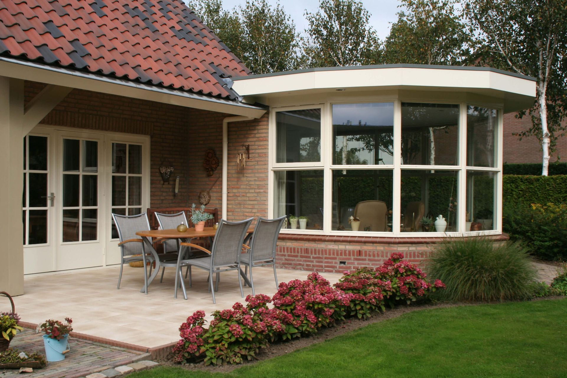 Patio outside a brick house with a round bay window, outdoor table and chairs, and flower beds.
