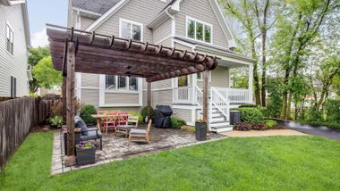 Backyard patio with wooden pergola, outdoor seating, stone pavers, and a green lawn beside a house