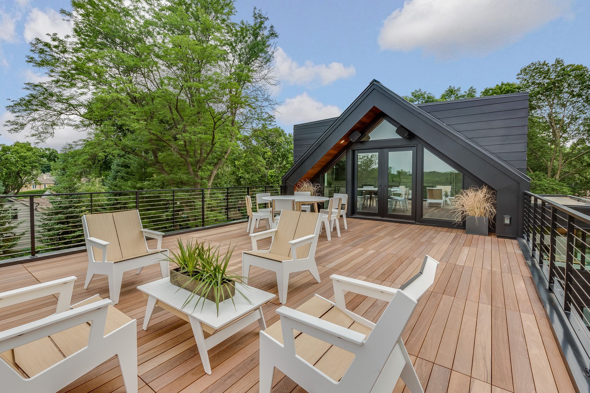 Rooftop deck with white chairs, wooden flooring, potted plants, and a modern black gabled house under a blue sky.