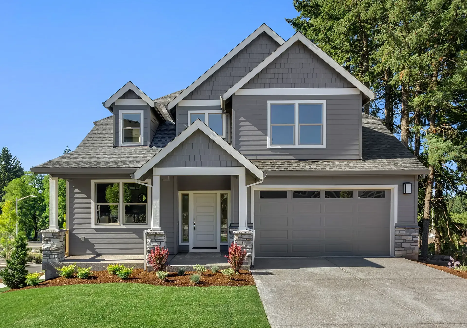 Gray two-story suburban house with a front porch, garage, and green lawn under a clear sky