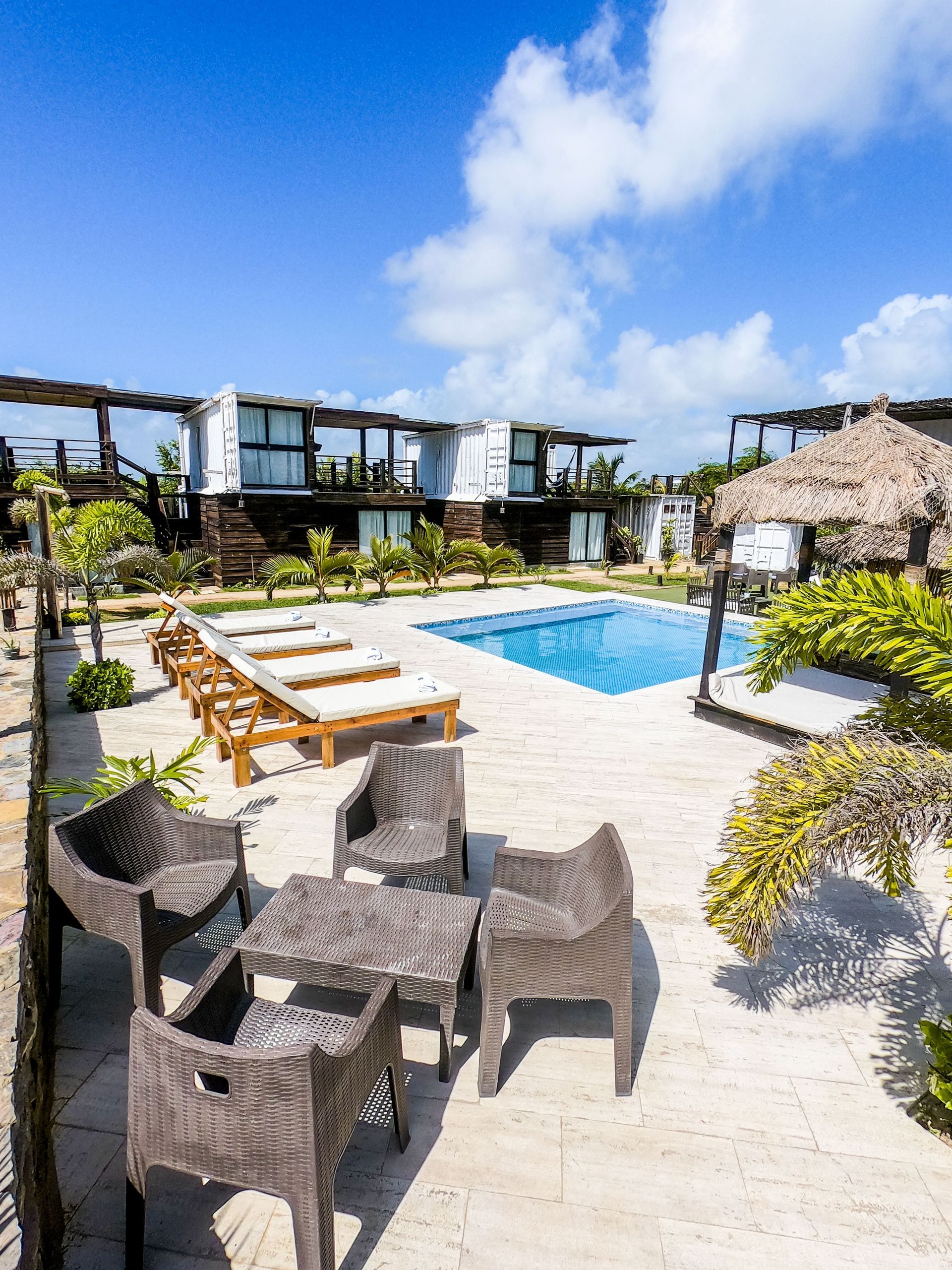 Resort pool with wicker chairs, lounge beds, and a pergola under a bright blue sky