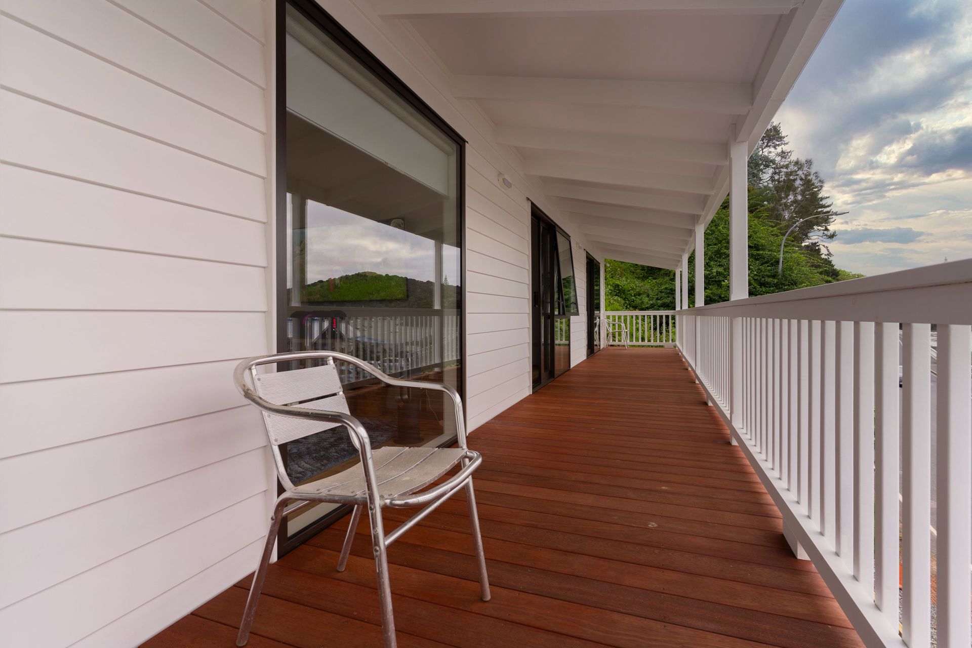 Covered porch with red wooden floor, white railing, and a metal chair beside a large window.