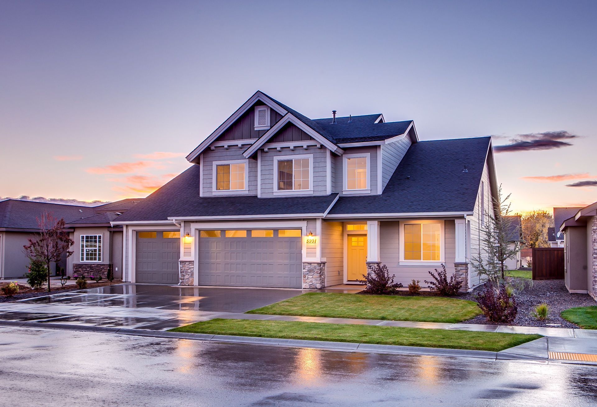 Suburban house at dusk with wet driveway, warm lights glowing, and a cloudy sunset sky