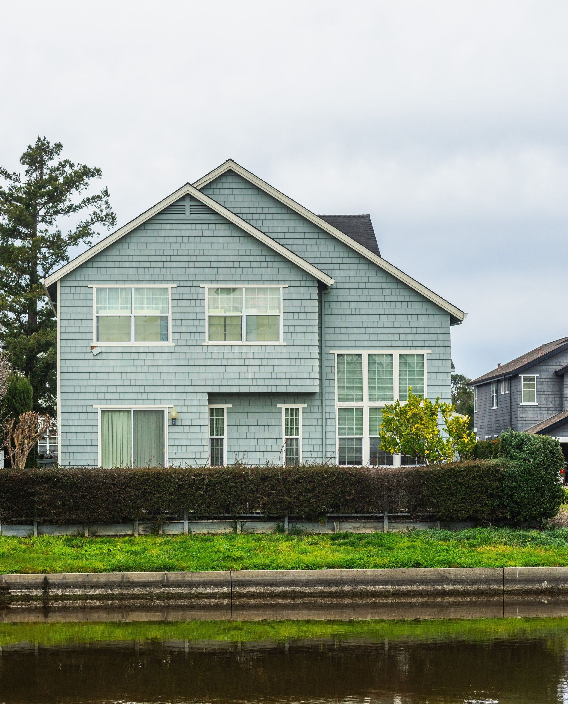 Gray two-story house with white trim behind a hedge, beside a canal under a cloudy sky