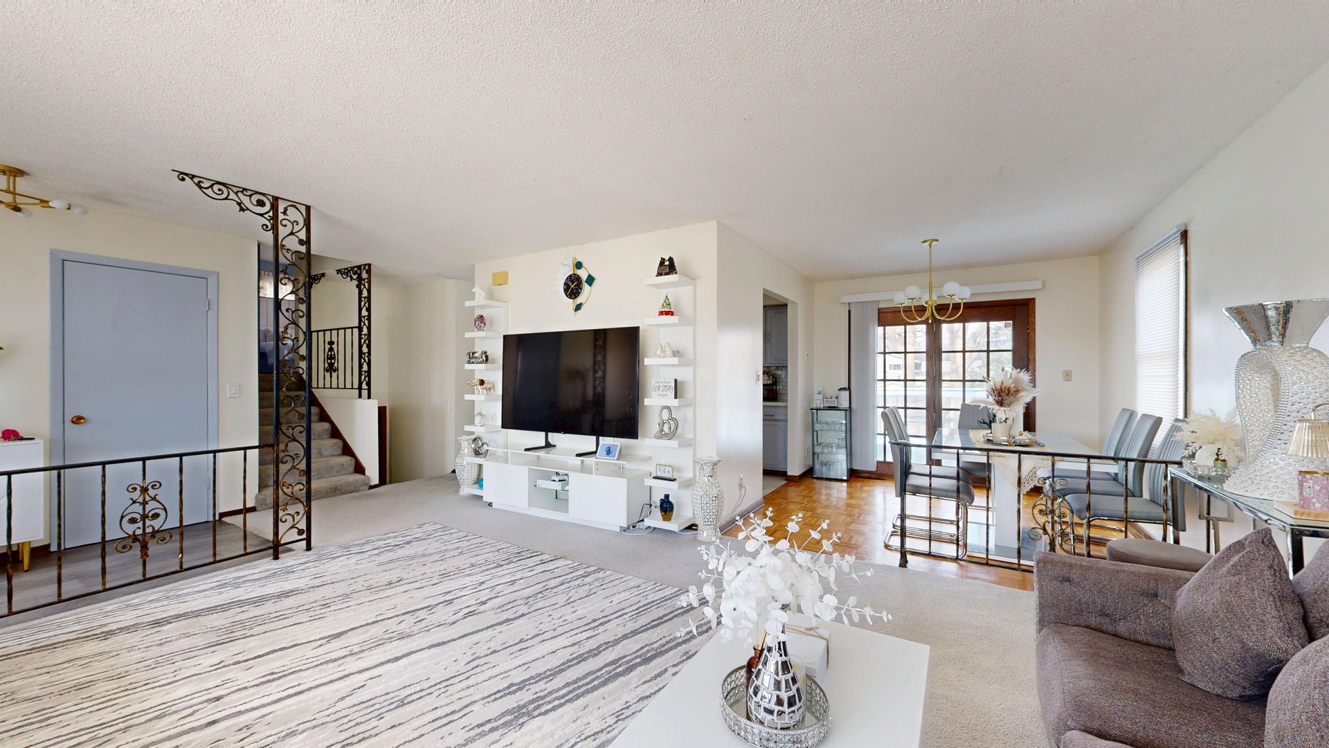 Bright open living room with white built-ins, TV, staircase, patterned rug, and gray sofa by glass doors