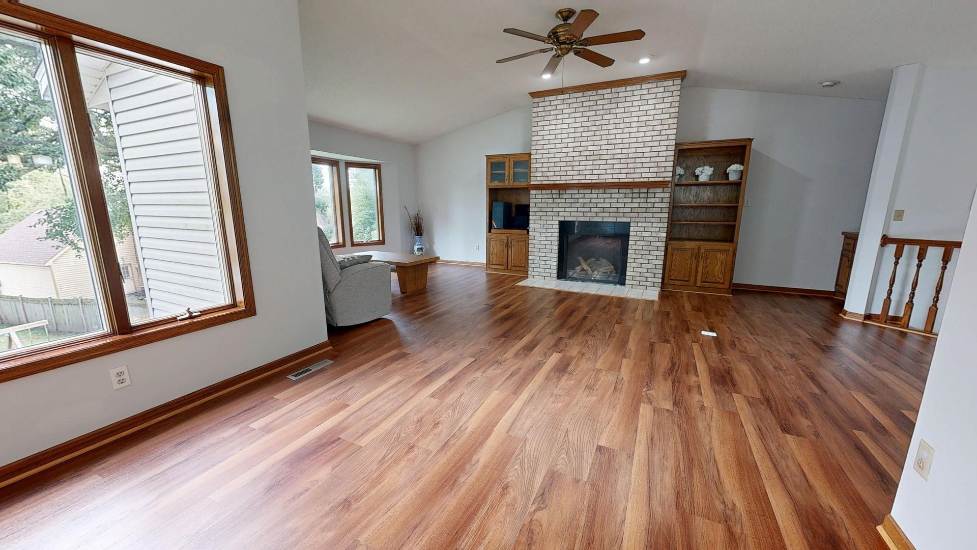 Empty living room with hardwood floors, stone fireplace, ceiling fan, and large windows