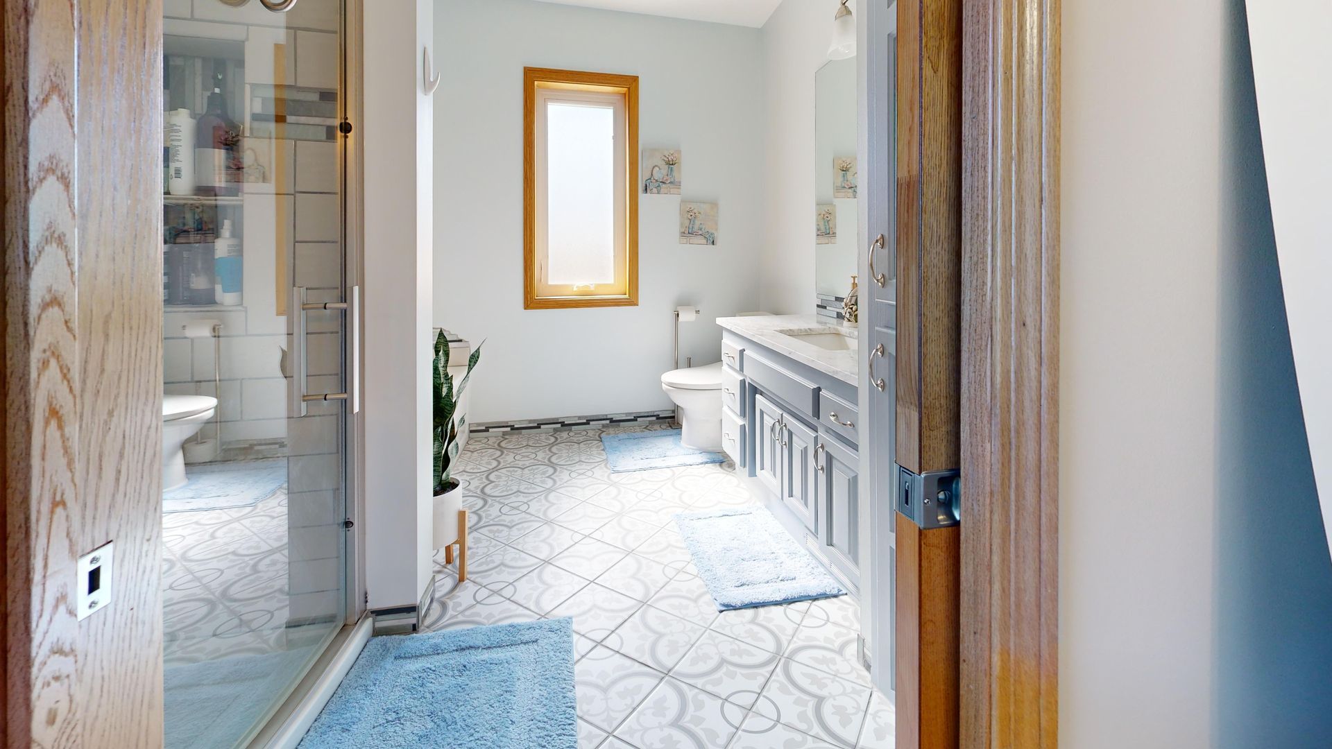 Bright bathroom with tiled floor, blue rug, mirror, and white vanity viewed through an open doorway
