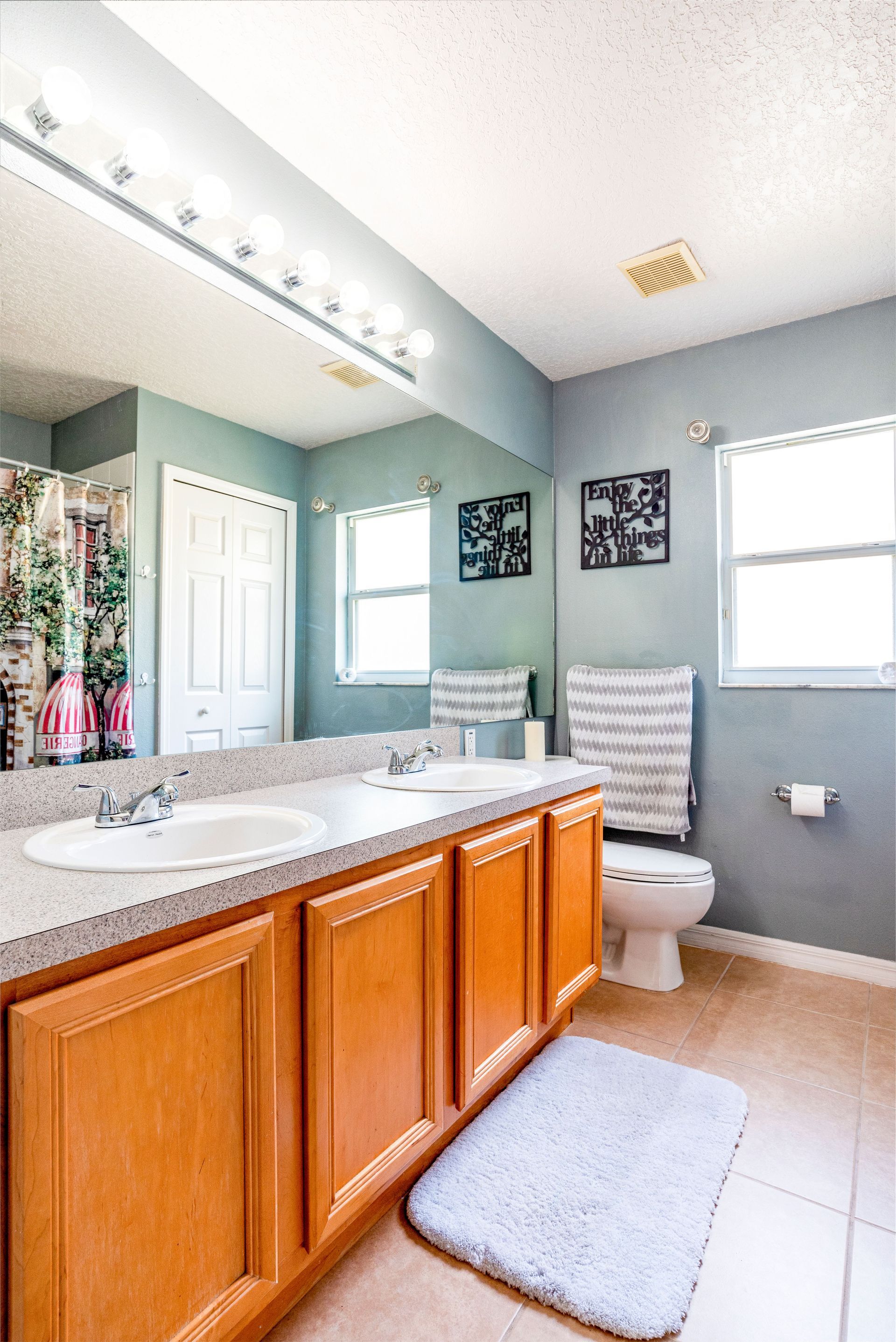 Bright bathroom with double vanity, large mirror, toilet, window, and gray walls.