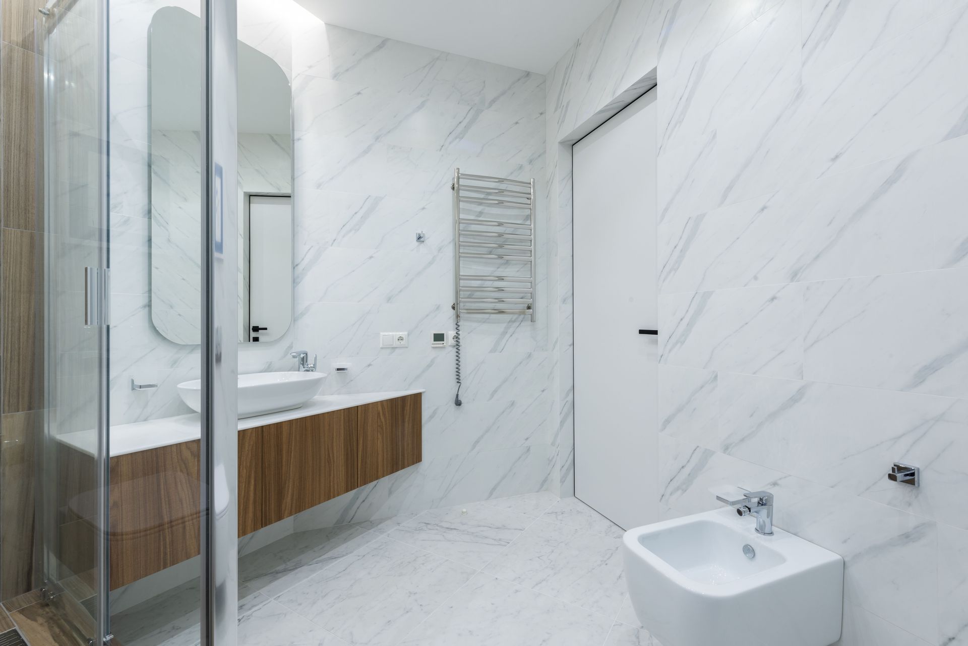 Modern white bathroom with marble walls, wood vanity, glass shower, and wall-mounted toilet.