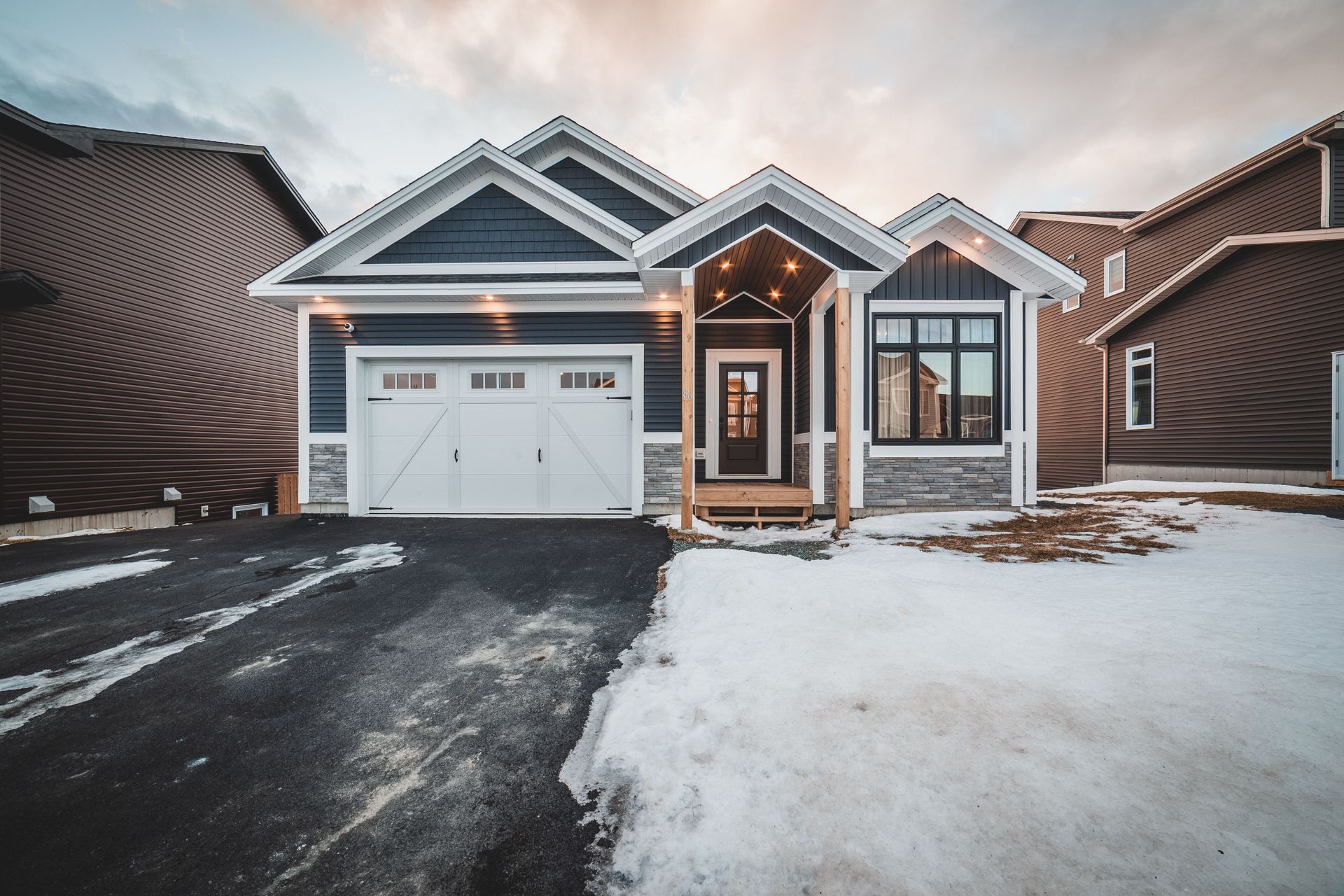 Snowy suburban house with a double garage, lit front entry, and dark driveway at dusk