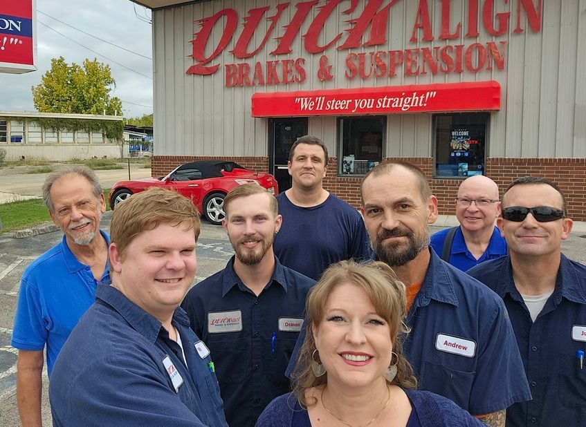 Seven people posing outside Quick Align Brake & Inspection shop with red signage and parked cars