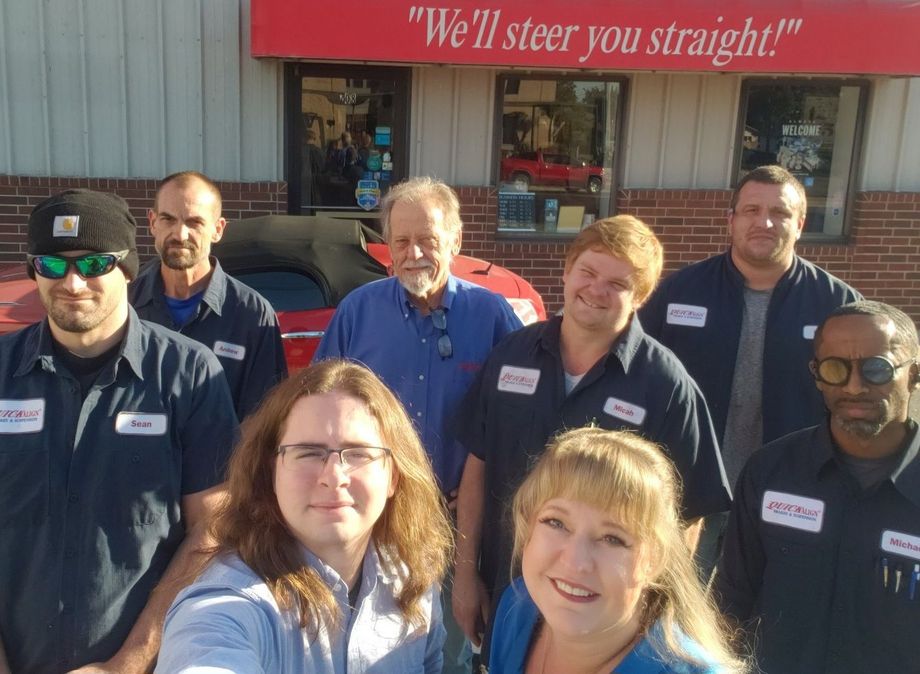 Seven people posing outside Quick Align Brake & Inspection shop with red signage and parked cars