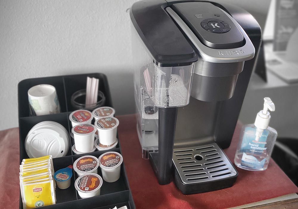 Coffee maker with K-cups, cups, and hand sanitizer on a wooden countertop