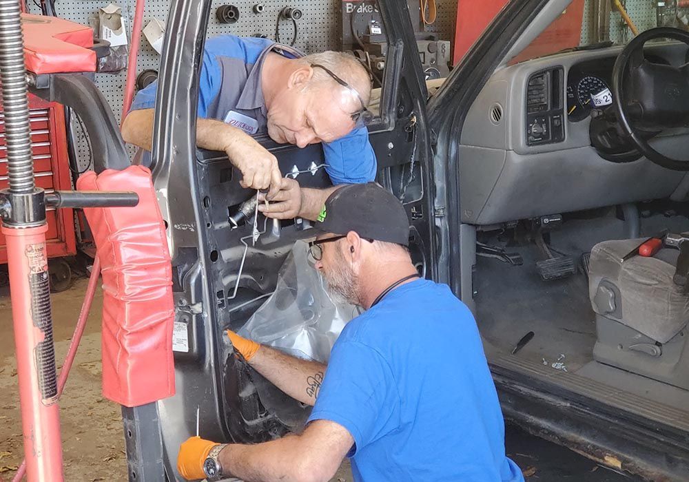 Two mechanics in blue shirts work inside a truck cab, inspecting wiring near the dashboard.