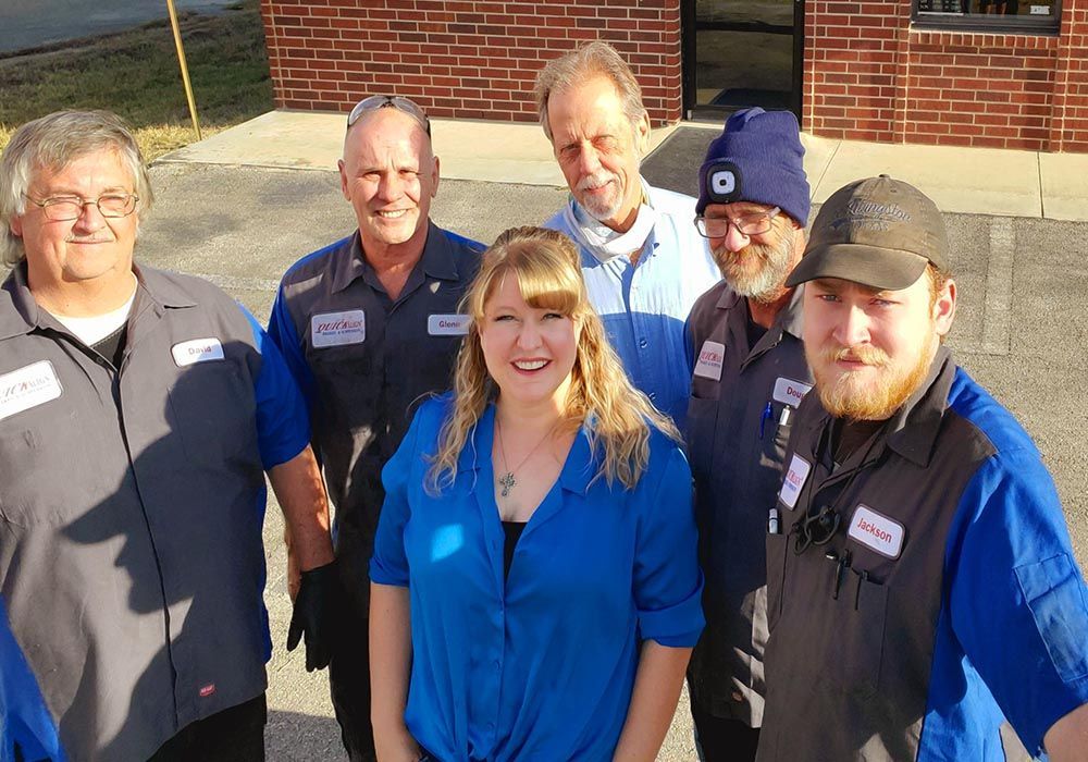 Five smiling people in work shirts standing outdoors in front of a brick building.