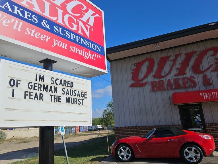 Red sports car parked outside a brake shop with a sign reading, “I’m scared of German sausage I fear the wurst.”