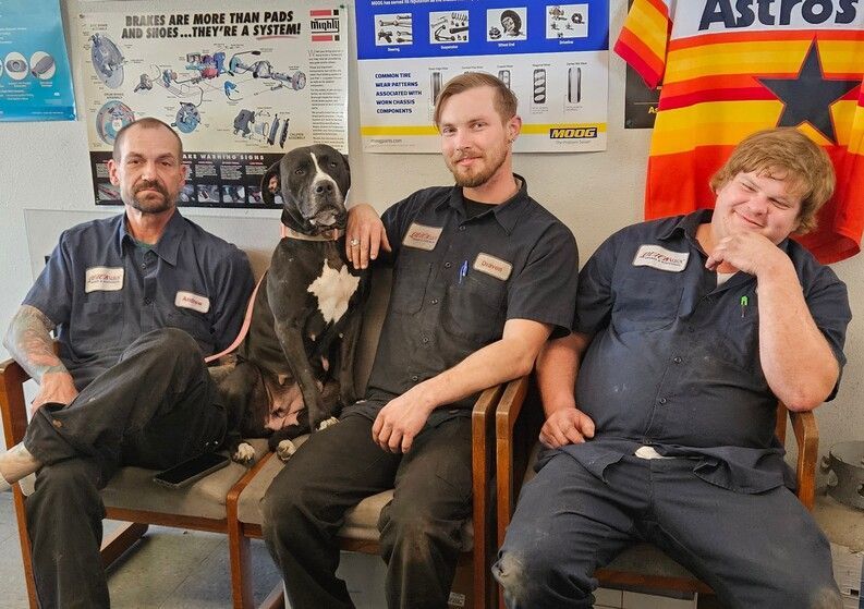 Three men in work uniforms sit with two dogs in an office, with posters and an Astros flag behind them.