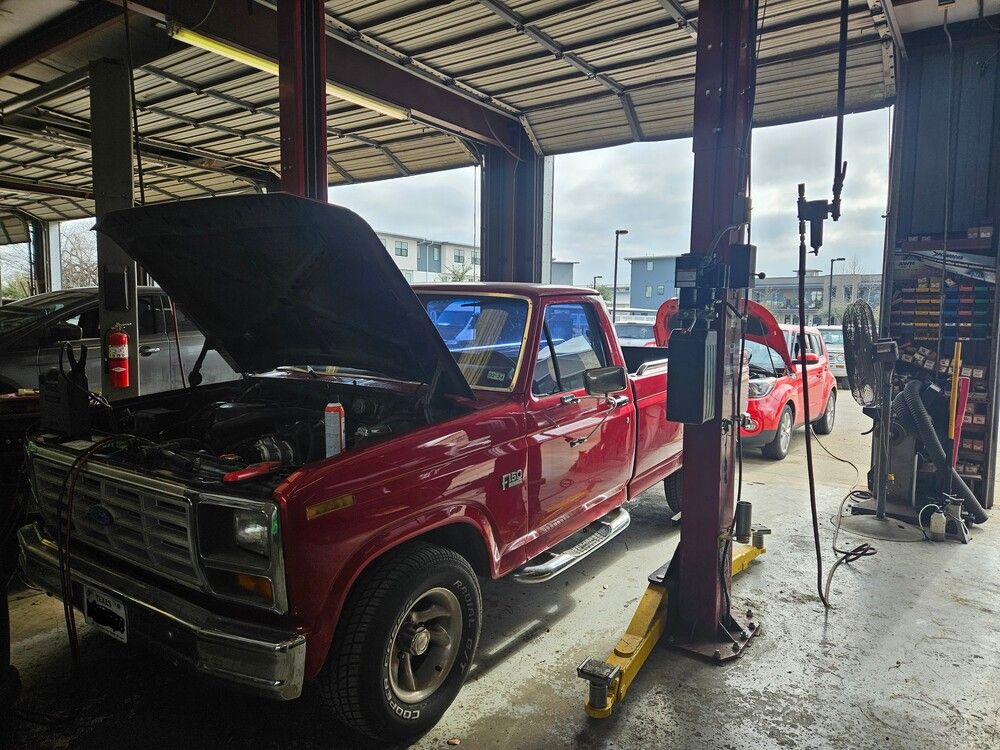 Red pickup truck with hood open in a garage, with a red car behind it and mechanic equipment nearby