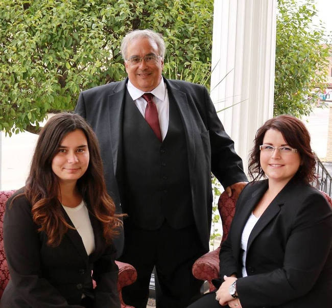 Three people in business attire posing outdoors on a porch, with greenery and white columns behind them.
