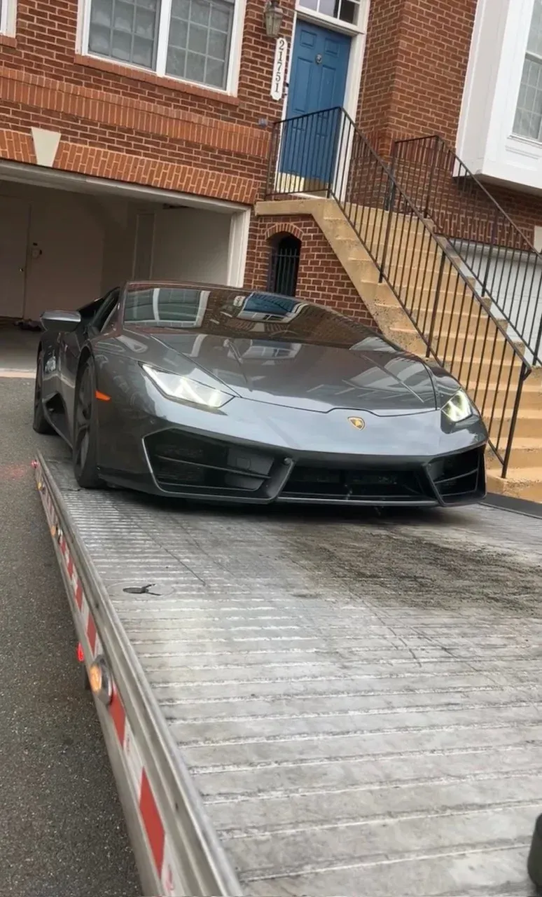 A gray Lamborghini sports car loaded onto a flatbed tow truck parked in front of a brick house.