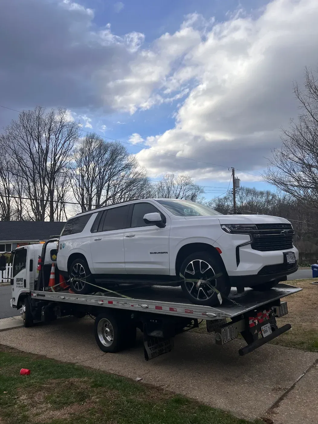 A white SUV being transported on the back of a flatbed tow truck on a street under a partly cloudy sky.