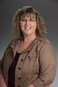 Woman with curly hair, wearing glasses, smiling, and a tan jacket.