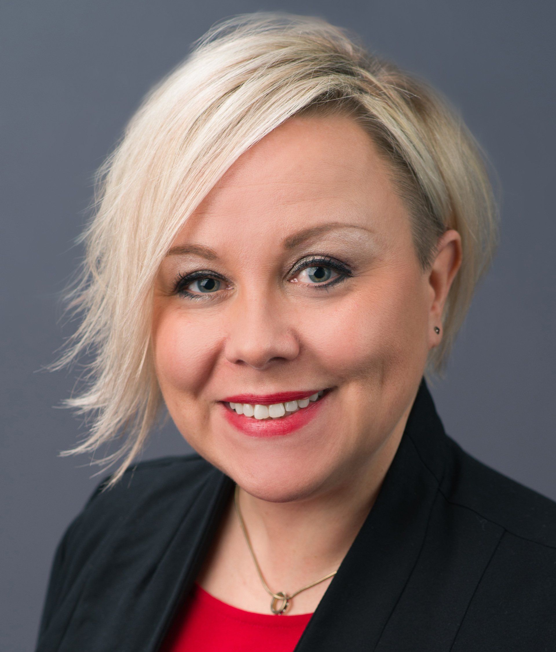 Blonde woman in black blazer smiles; red top, gray background.