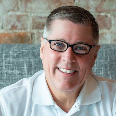 Man in a white shirt and glasses smiling, seated near a brick wall and a grey cushion.