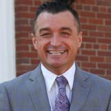Smiling man in a gray suit and purple tie, standing outside a brick building.