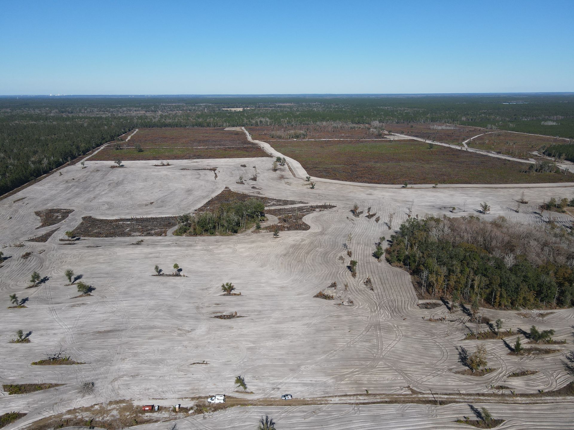Aerial view of a large, cleared plot of land with patches of brown soil and remaining forest vegetation under a clear sky.
