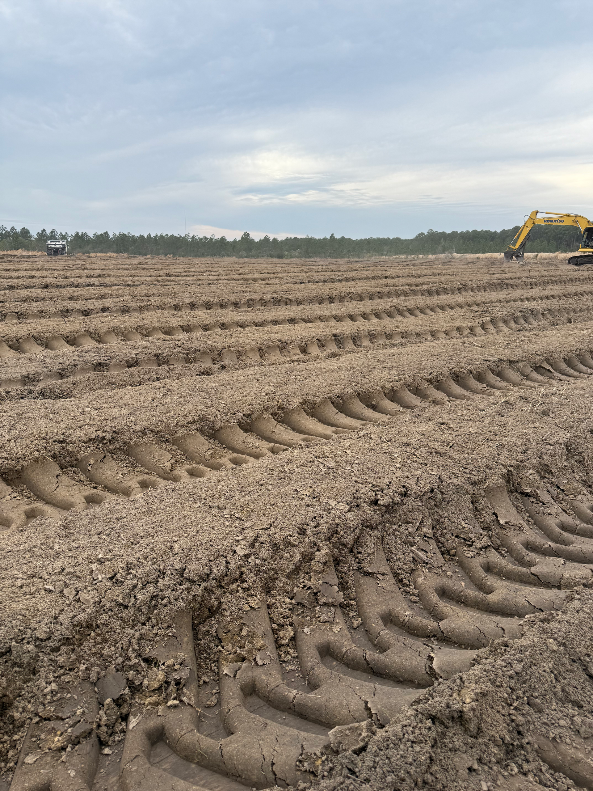 A yellow excavator works on a vast field of dark soil with deep, parallel tire tracks under a cloudy sky.