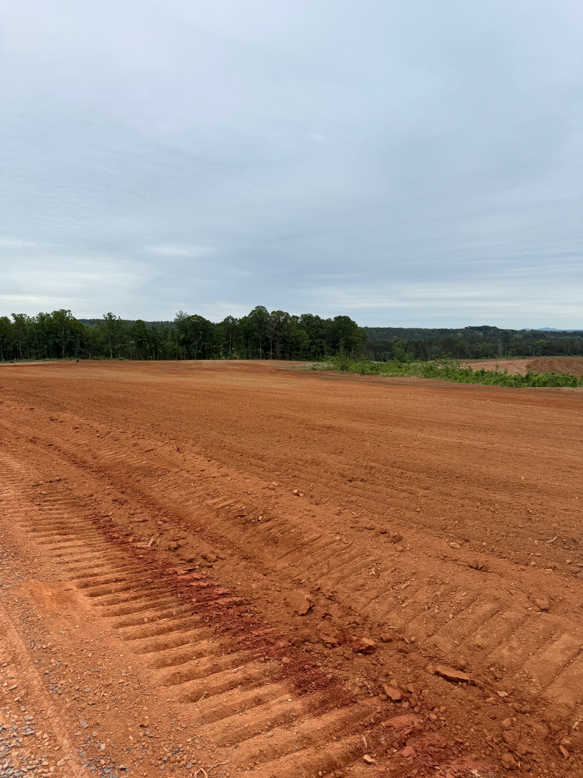 A field of red, freshly tilled soil under an overcast sky, with a line of trees in the background.