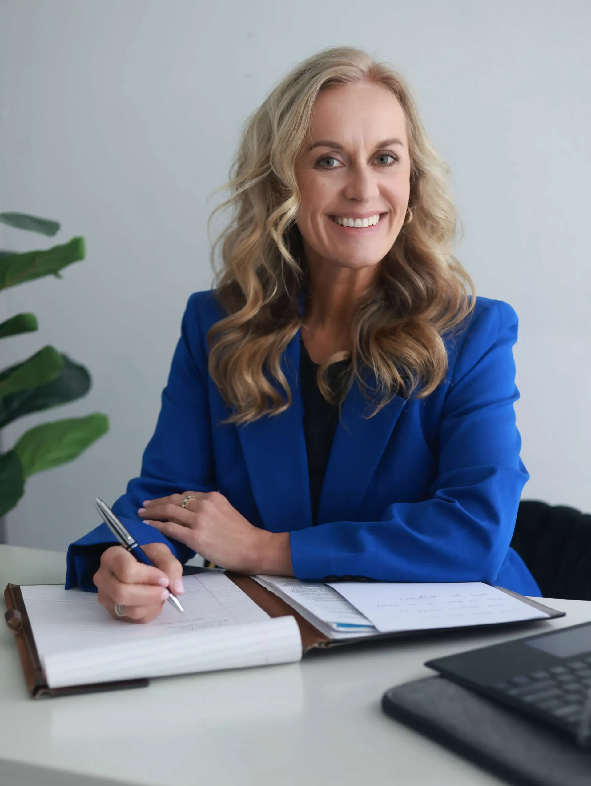 A woman in a blue jacket is sitting at a desk writing in a notebook.