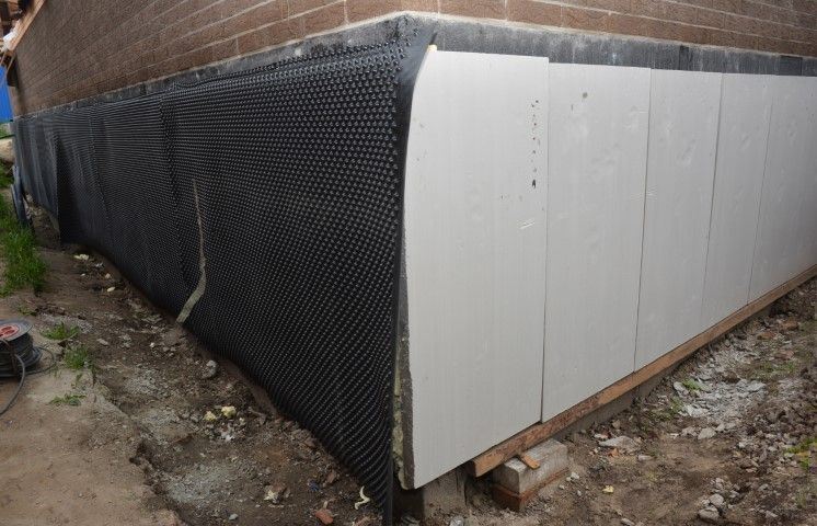 Exterior building wall covered in black waterproofing and white siding panels along a brick foundation