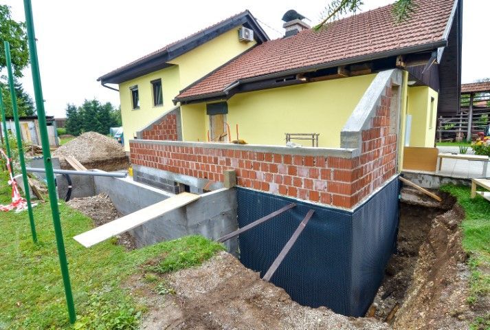 House under construction with brick foundation walls, dark waterproofing, and concrete supports beside a yellow home