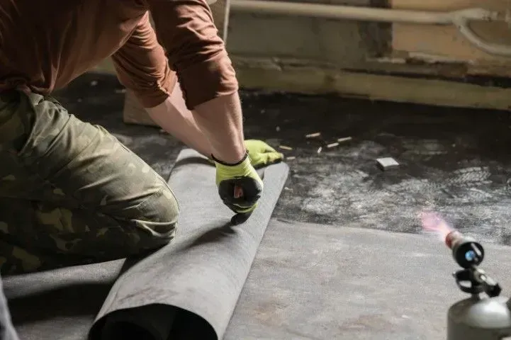Person rolling out black flooring material on a concrete floor with a tape measure and gloves