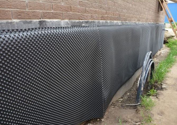 Black dimpled waterproofing membrane along a brick foundation wall beside a house