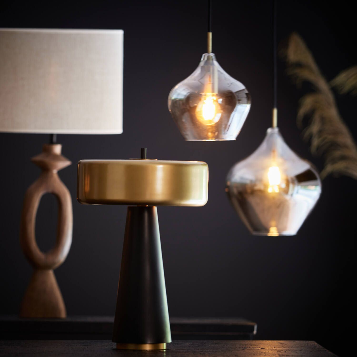 Lampen op een donkere tafel: een houten tafellamp, een goud-zwarte bureaulamp en twee glazen hanglampen.
