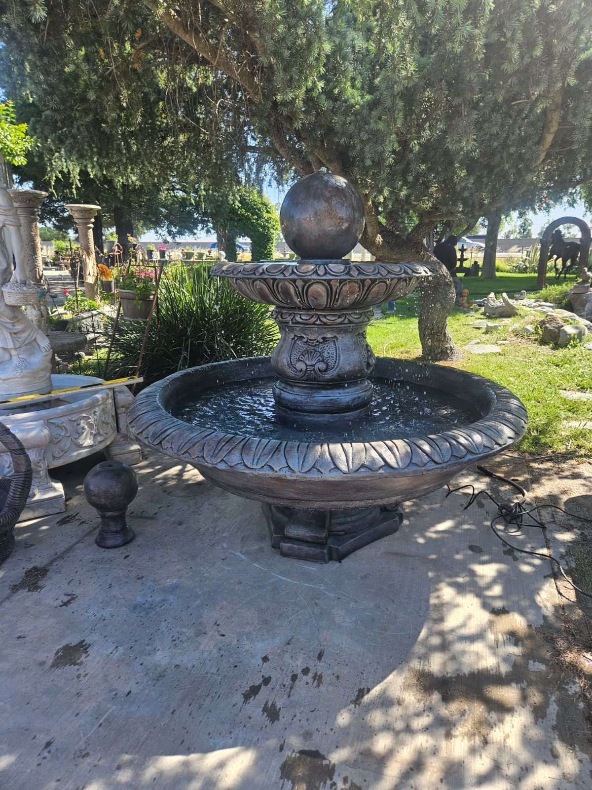 Stone fountain with two-tier basin in a shady garden, surrounded by trees and benches.