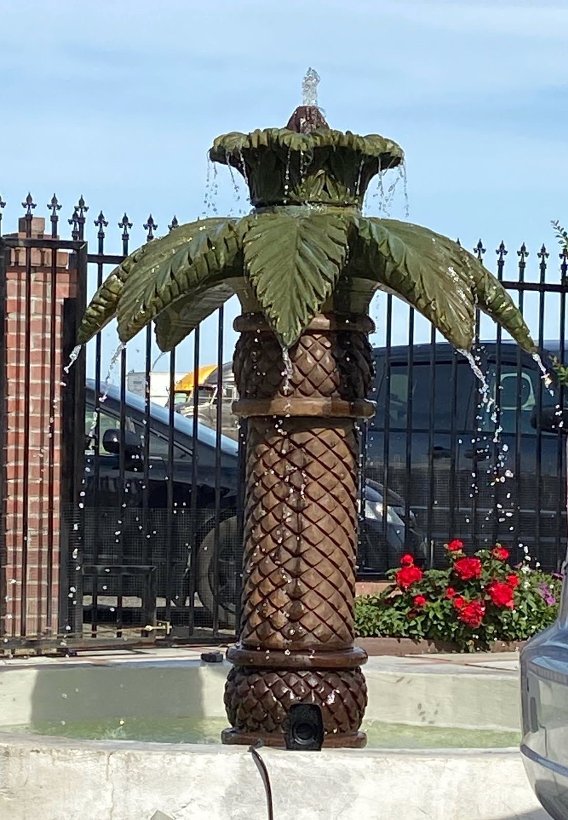 Decorative palm-tree fountain sculpture beside a black iron fence, with red flowers nearby.