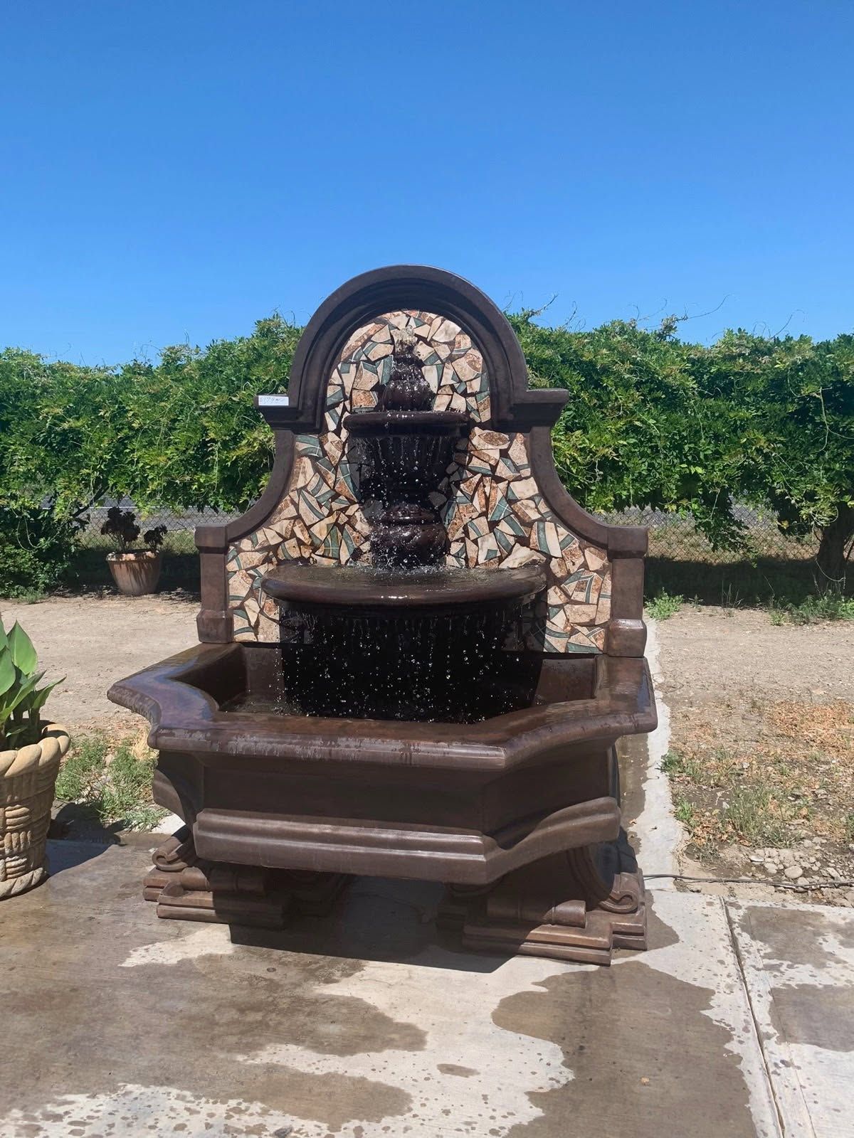 Stone fountain with a decorative arched back and water spilling into a basin, outdoors in a vineyard.