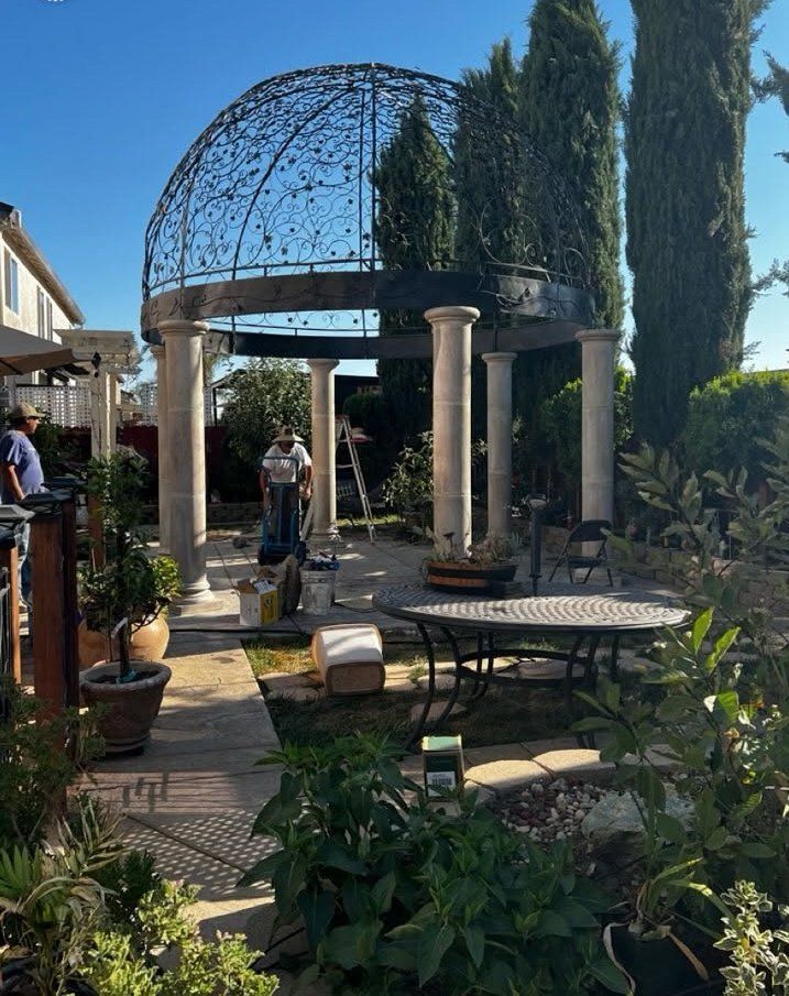 Garden patio with a vine-covered gazebo, columns, tables, and tall cypress trees in sunlight