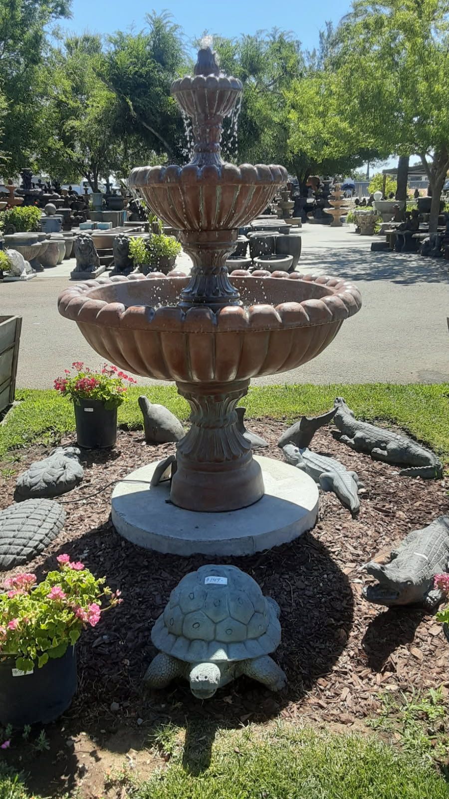 Tiered stone fountain in a sunny garden, surrounded by rocks, flowers, and trees.