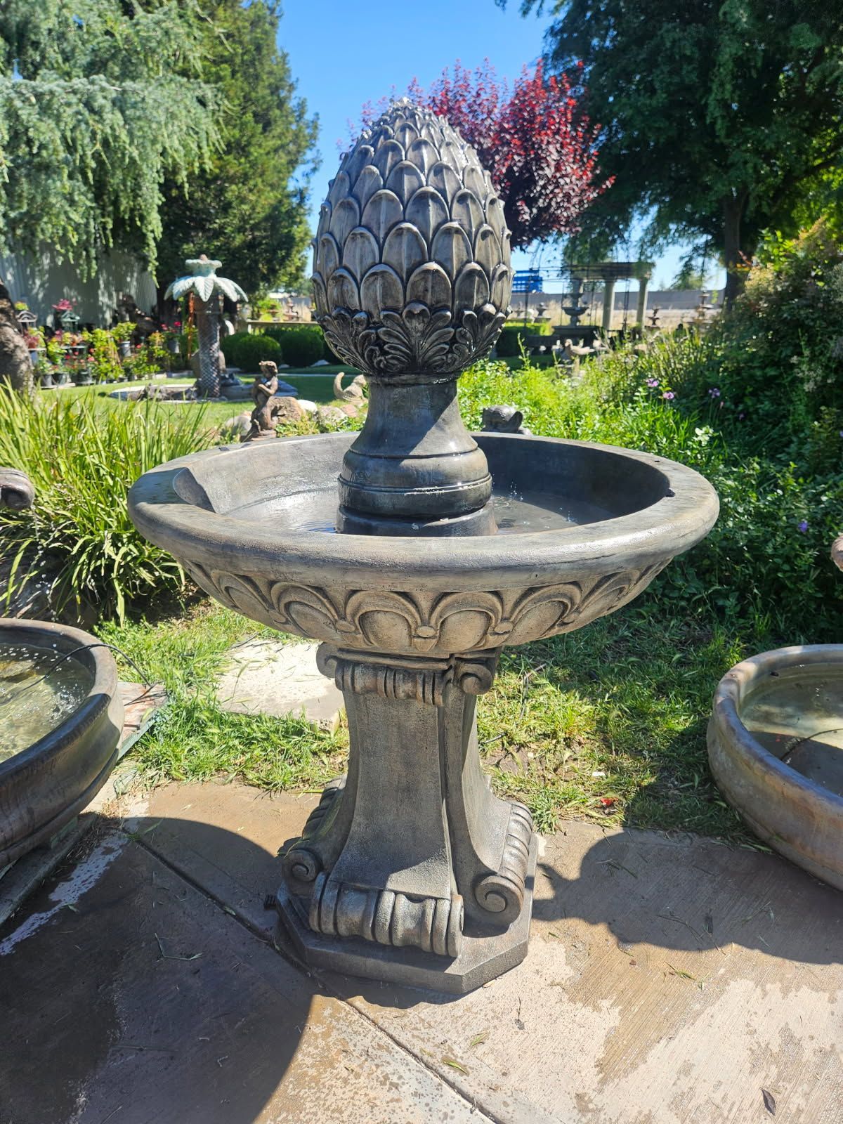 Stone fountain with pineapple finial in a sunny garden, surrounded by flowers and greenery