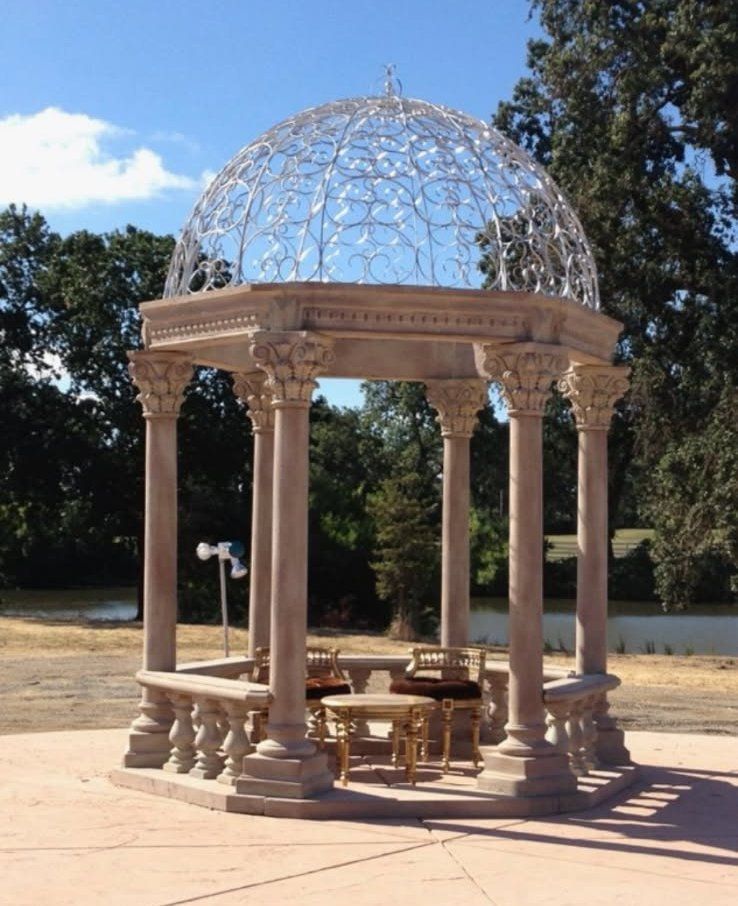 Ornate gazebo with domed lattice roof and stone columns in a sunny park.